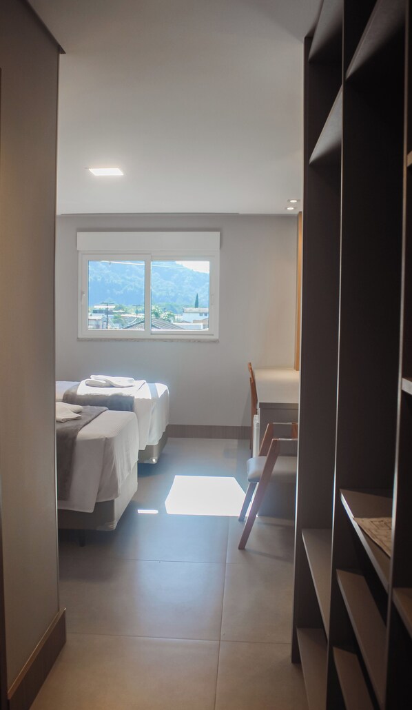 Un pasillo estrecho con una cama, un escritorio y una ventana con vistas a edificios y montañas.