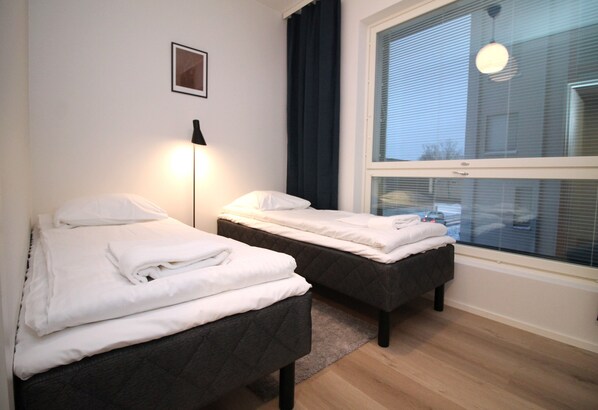 A hotel room with two beds, a nightstand, a lamp, a window with blinds, and a framed picture on the wall.