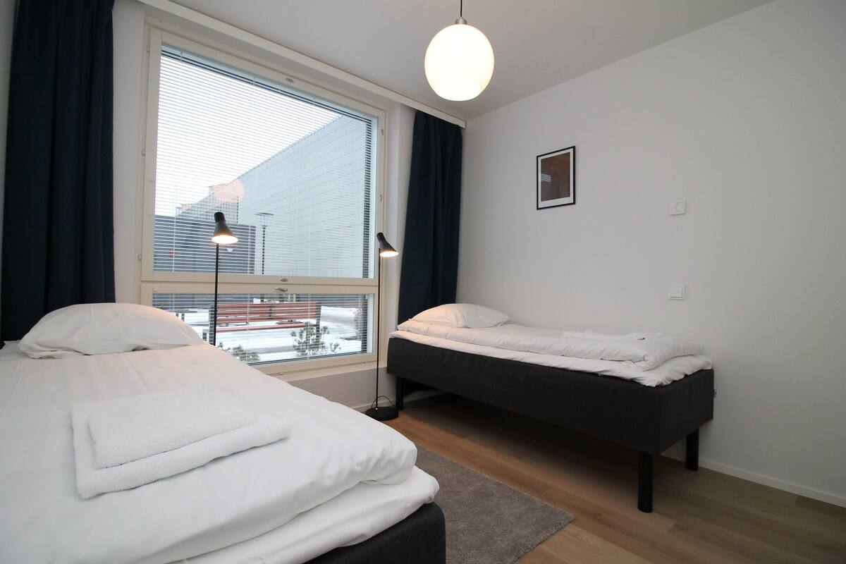 Ein modernes Schlafzimmer mit zwei Betten, einem großen Fenster mit Jalousien, einer Pendelleuchte und einem kleinen Bild an der Wand.