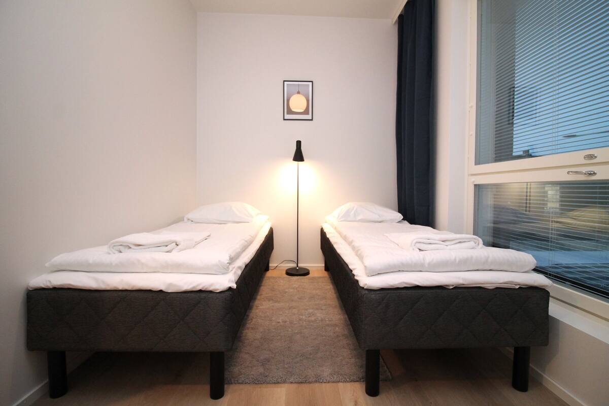 Ein kleines, sauberes Zimmer mit zwei Einzelbetten, einer Stehlampe und einem Fenster mit Jalousien.