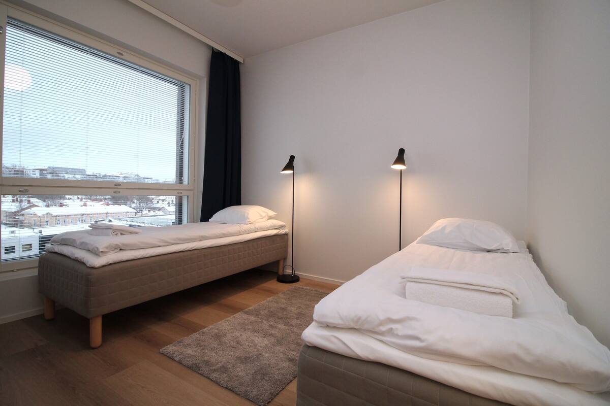 Ein Schlafzimmer mit zwei Betten, einem Fenster mit Jalousien, einer Stehlampe und Blick auf Gebäude.