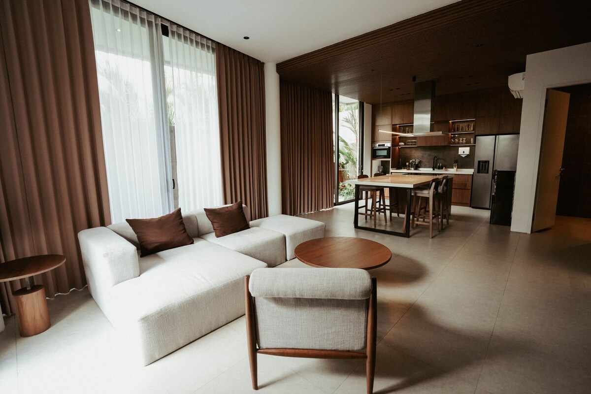 Un salon moderne avec un canapé, un fauteuil et une table basse ronde. On aperçoit à l’arrière-plan un coin cuisine avec une table à manger et des chaises.