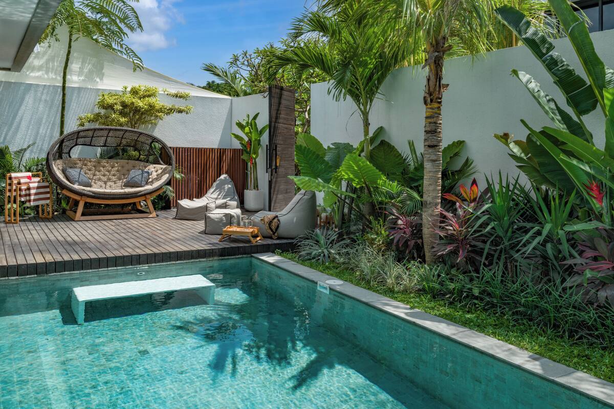 Ein Poolbereich mit Holzterrasse, gepolsterten Sitzgelegenheiten und tropischen Pflanzen.
