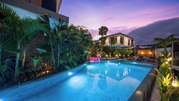 Una piscina con un flotador rosa con forma de flamenco, rodeada de exuberante vegetación tropical y un edificio moderno al fondo.