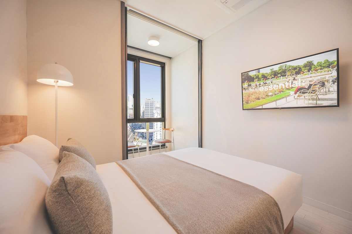 Une chambre d’hôtel avec un lit, une table de chevet, une lampe et une grande fenêtre donnant sur la ville.