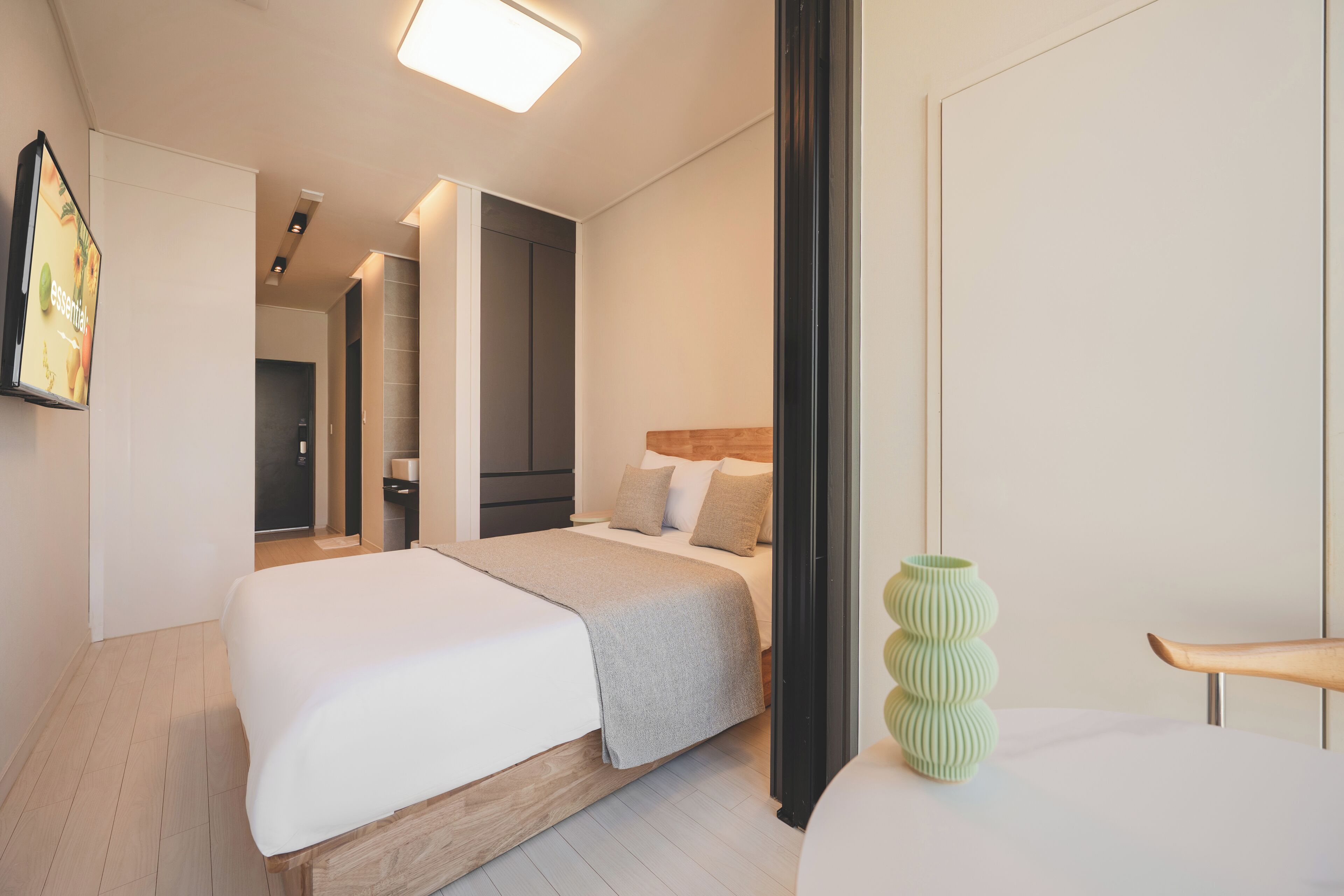 A modern hotel room with a large bed, a bedside table, a green vase, and a wooden chair.