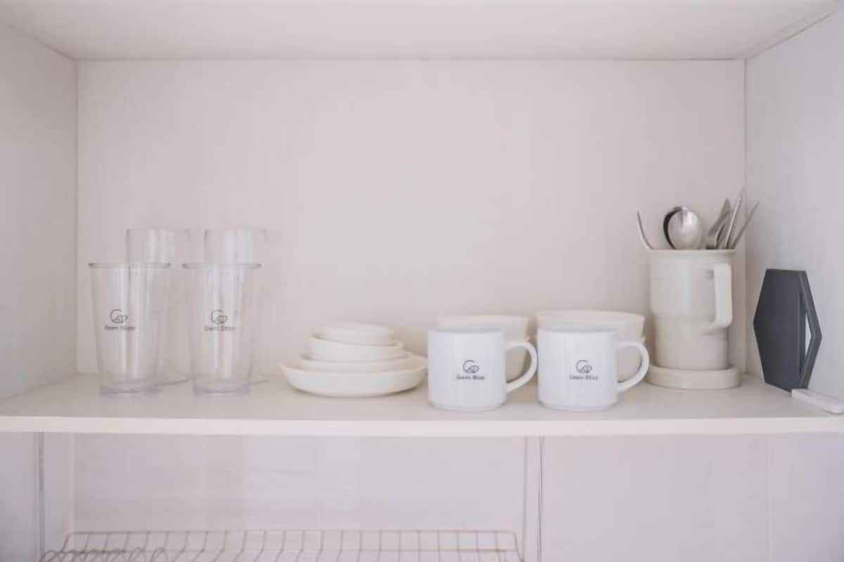Une étagère blanche avec des verres transparents, des assiettes blanches, des tasses blanches, une machine à café blanche et un haut-parleur blanc.
