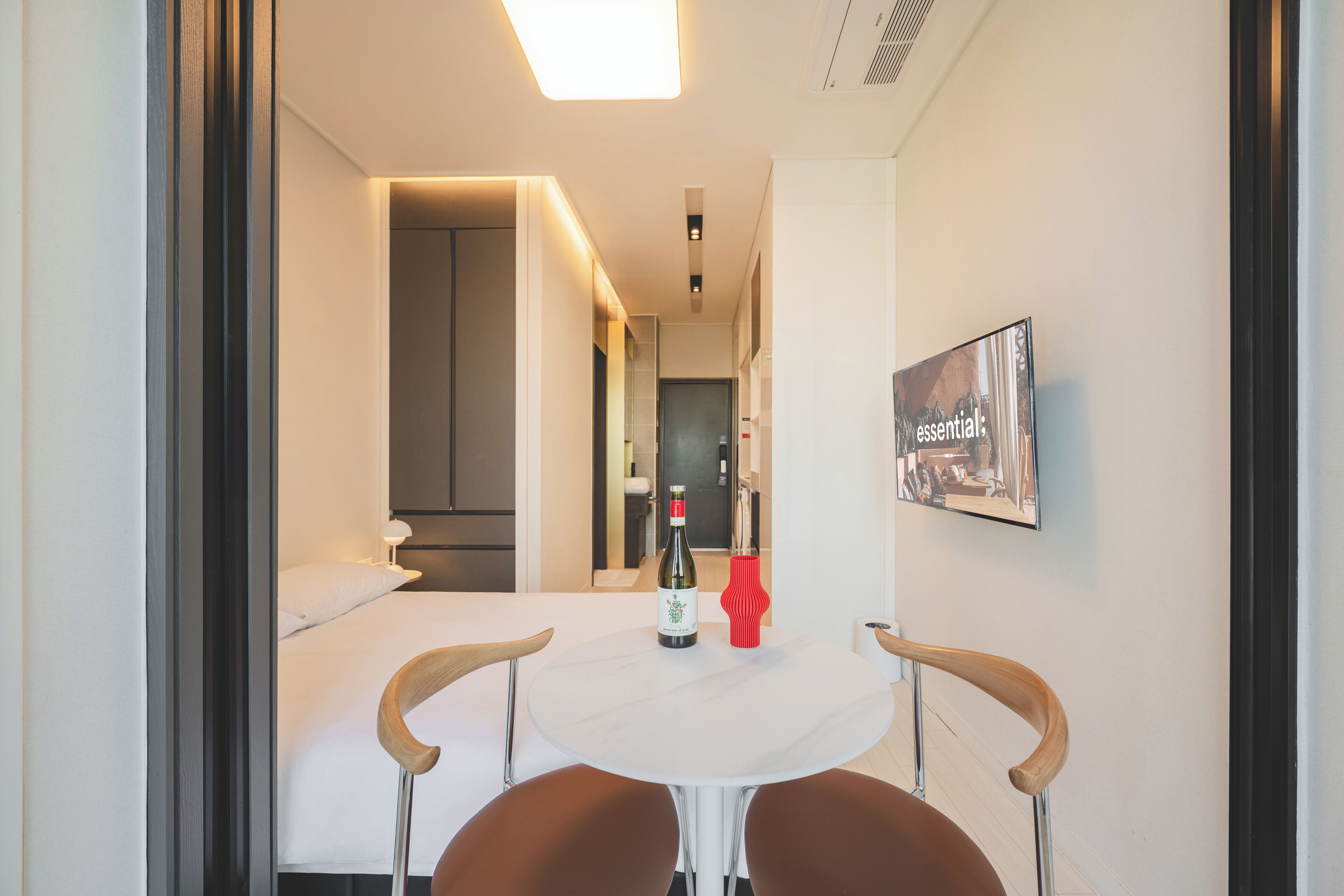 A hotel room with a bed, a small table with a bottle and a red vase, and a framed picture on the wall.