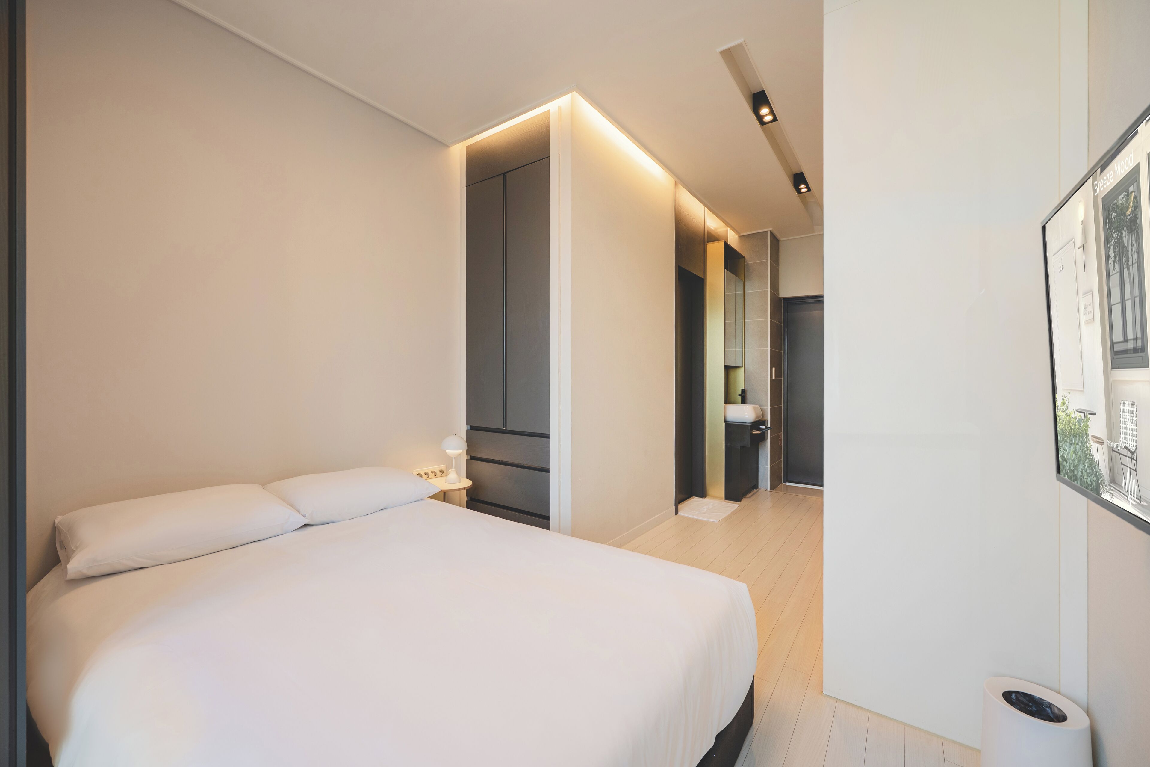 A modern hotel room with a large bed, a bedside table with a lamp, a bathroom with a mirror, and a framed picture on the wall.