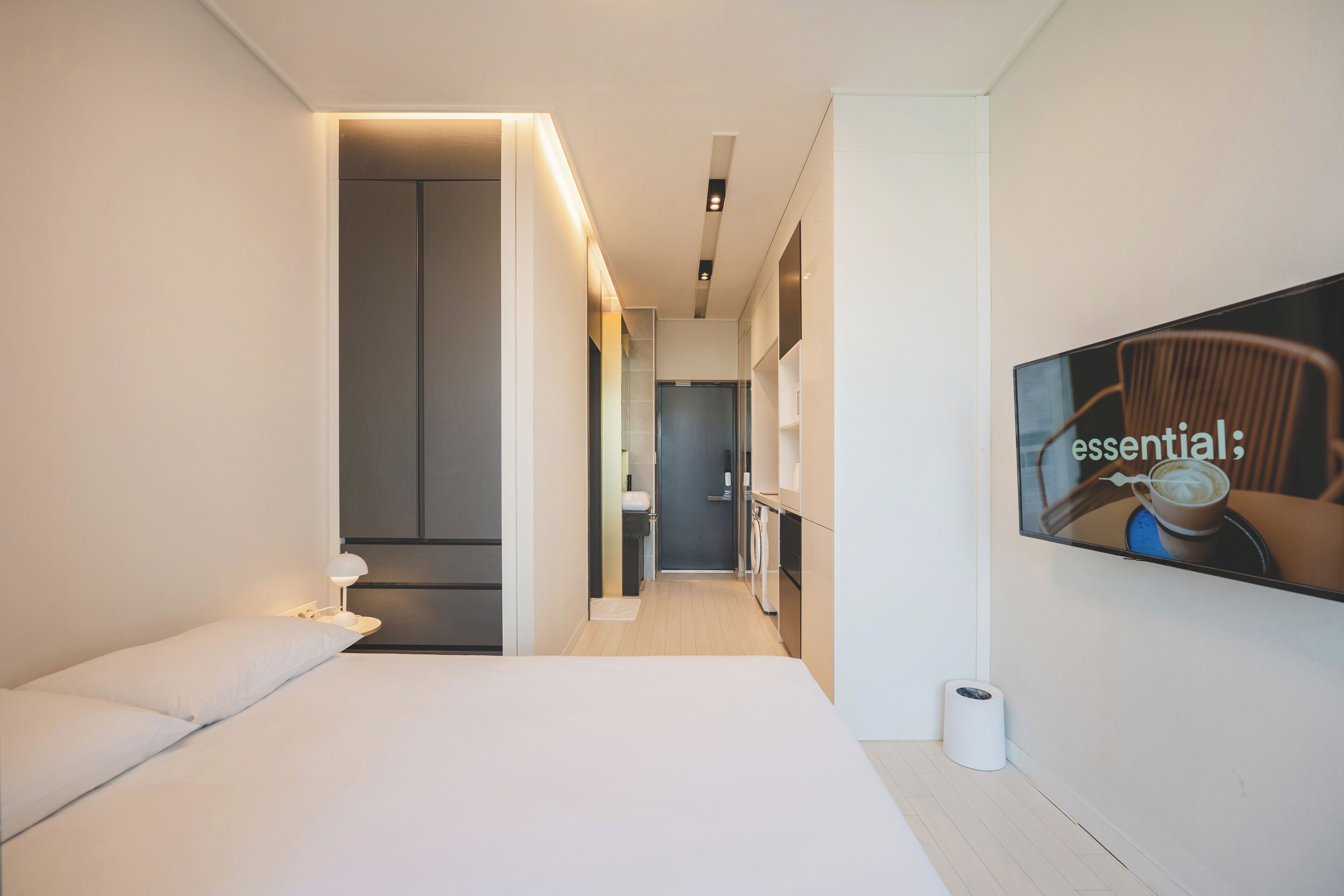 A modern hotel room with a bed, a TV mounted on the wall, and a hallway leading to a bathroom.