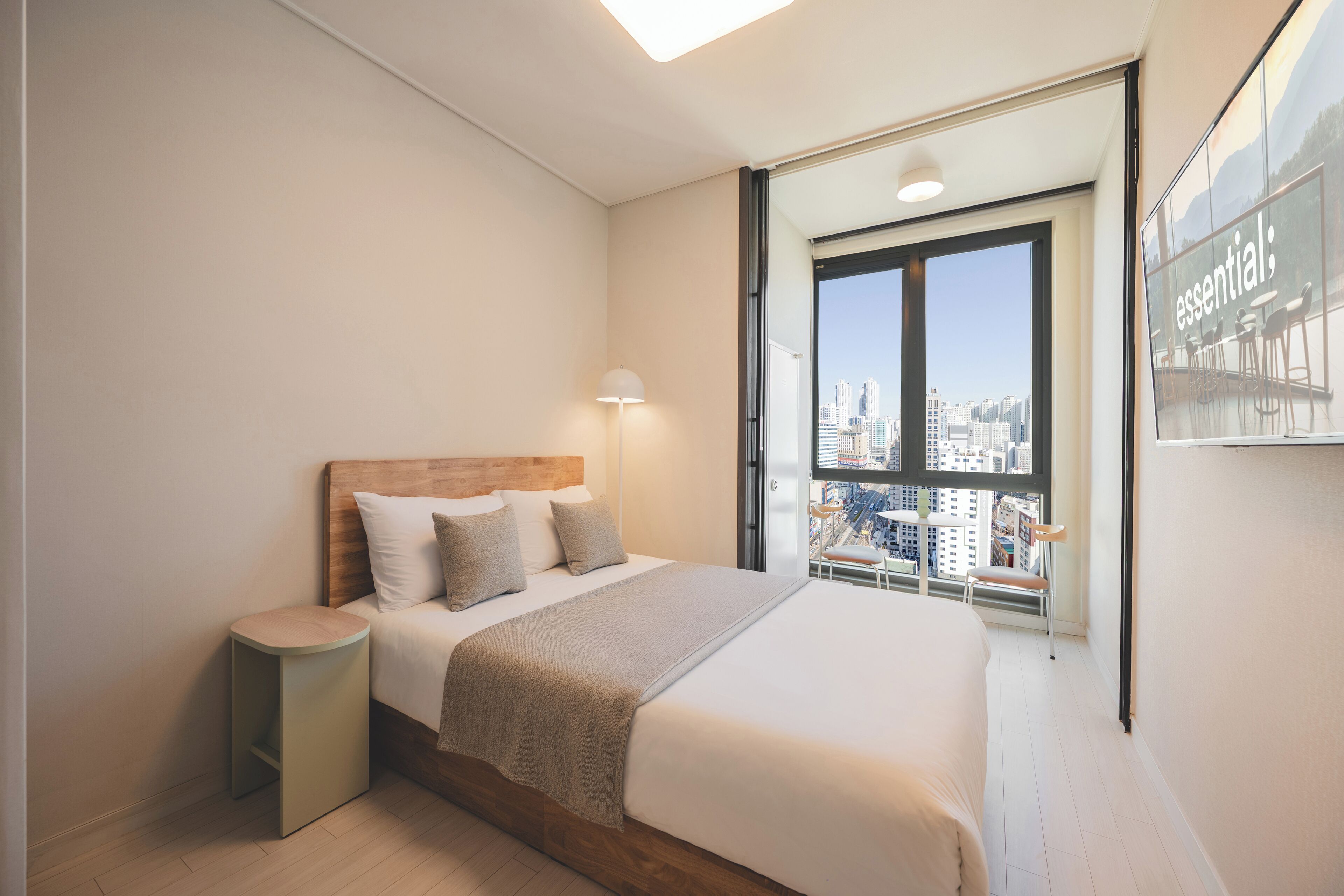 Habitación de hotel moderna con una cama grande, una mesita de noche, un ventanal con vista a la ciudad y un cuadro enmarcado en la pared.