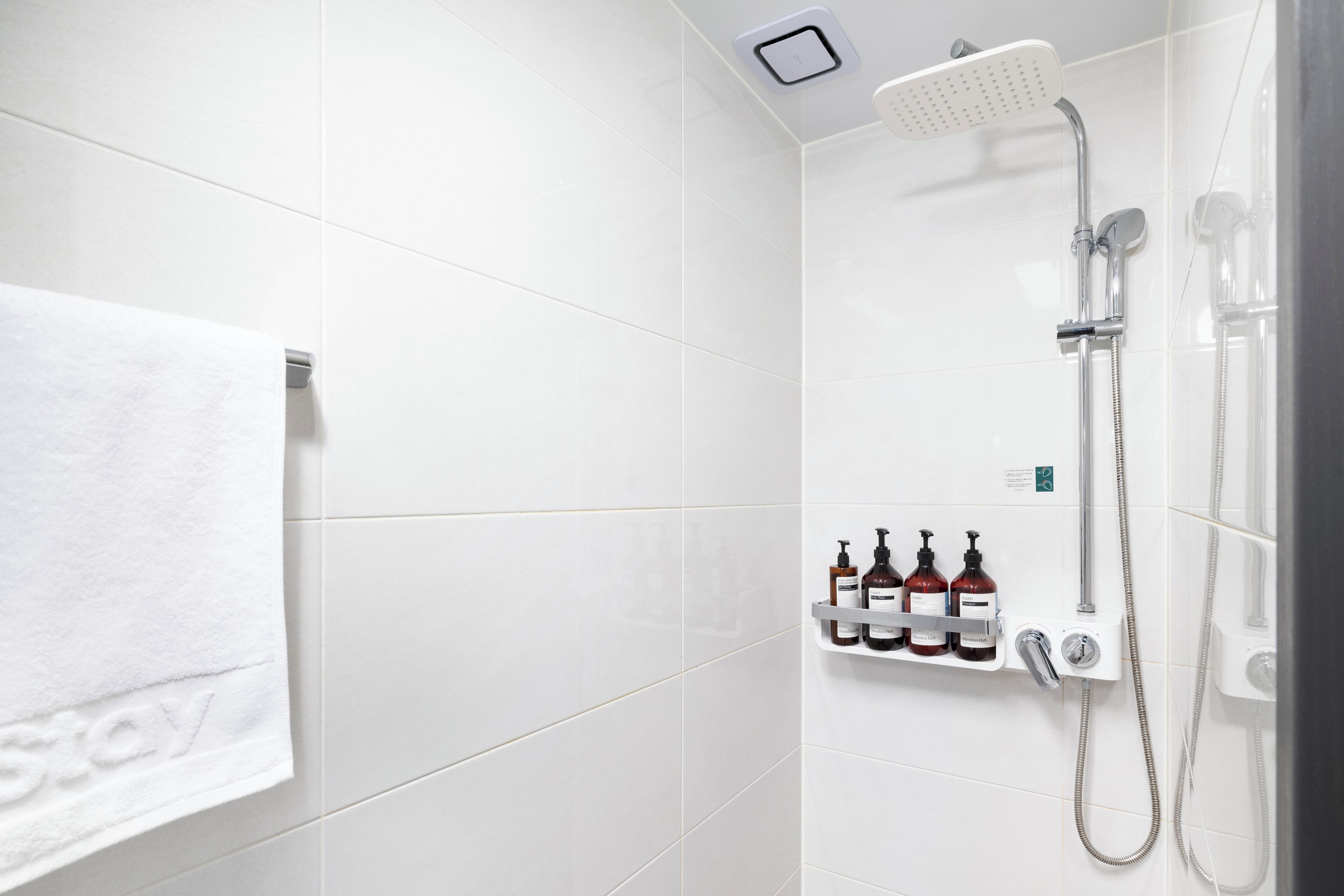 A modern bathroom with white tiles, a shower with a handheld showerhead, and a shelf with toiletries.
