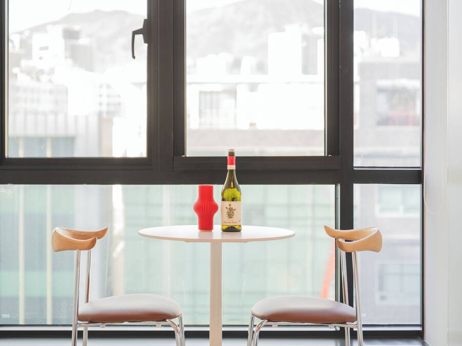 A room with a round table, two chairs, a bottle of wine, and a red vase.