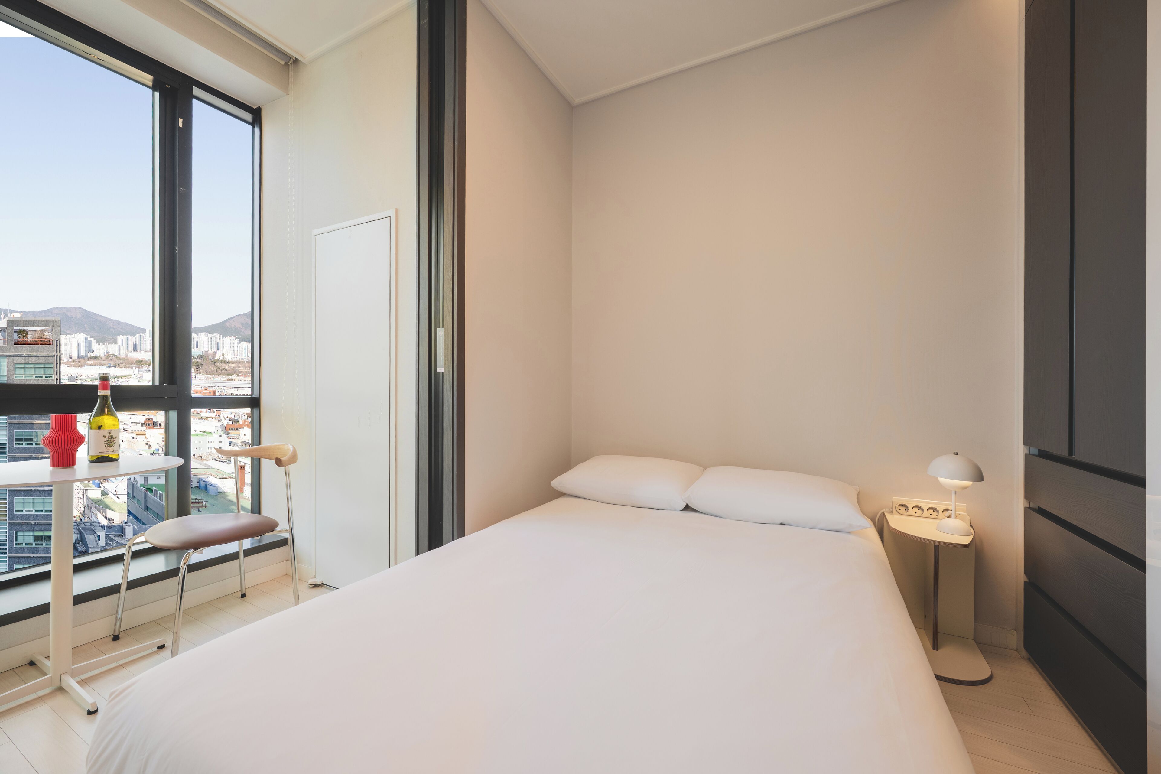 Een moderne slaapkamer met een groot bed, een klein tafeltje met een lamp, een stoel en uitzicht op een stadsgezicht door een groot raam.