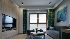 A modern living room with a flat-screen TV, a sofa, a coffee table, and a large window with curtains.
