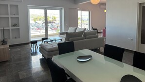 Un soggiorno moderno con divano, tavolo da pranzo e sedie. Grandi finestre offrono vista su un parcheggio e su edifici.