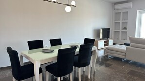 Zona pranzo moderna con tavolo bianco, sedie nere e lampada a sospensione. Si nota una TV e un divano sullo sfondo.