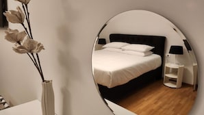 Una camera da letto con uno specchio rotondo, un letto con biancheria bianca, un comodino con una lampada e un vaso con fiori.
