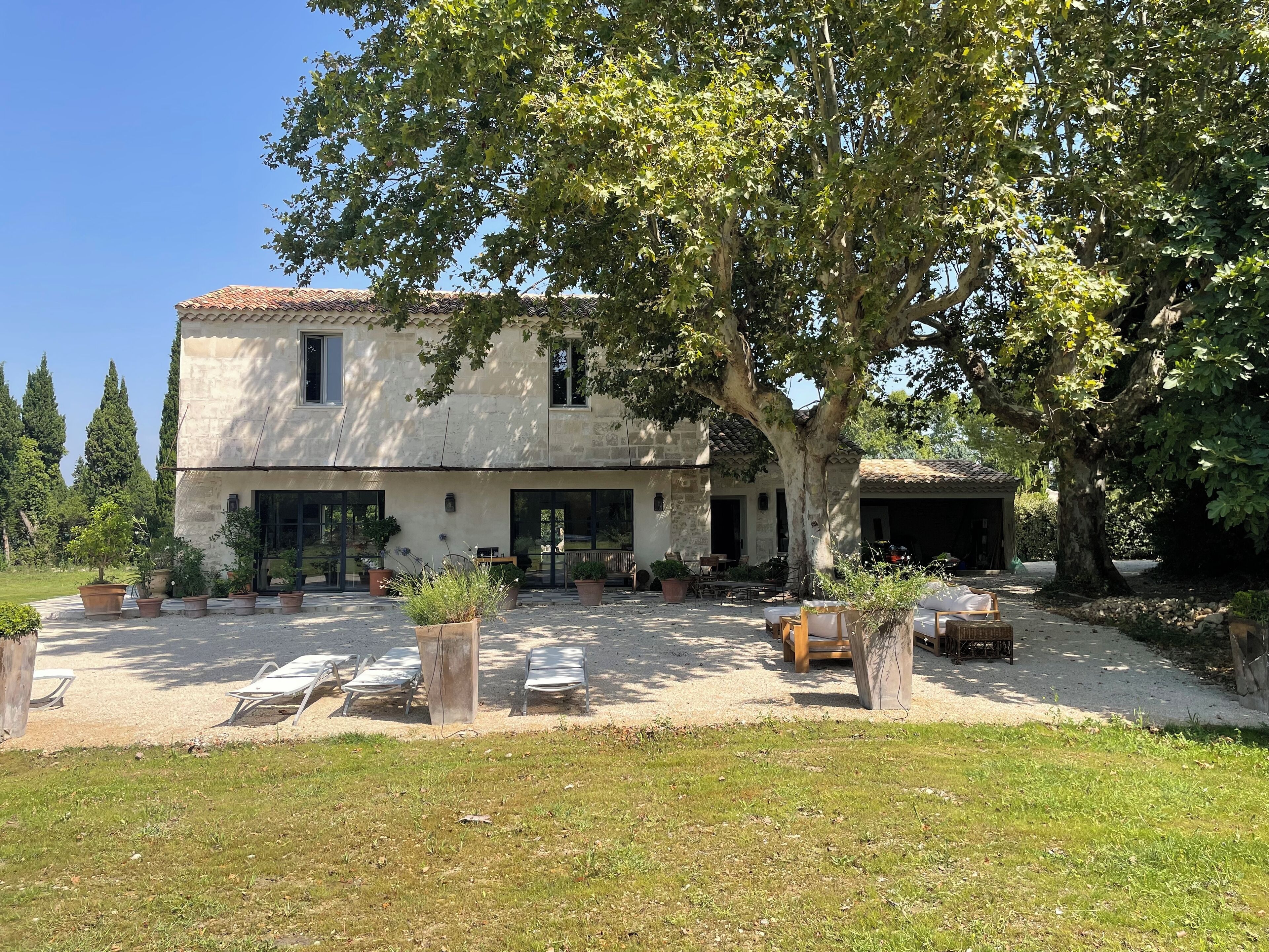 A two-story stone building with a tiled roof, a large tree in the front yard, and outdoor seating area with lounge chairs and tables.