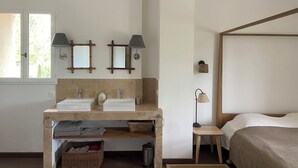 A bedroom with a bed, a bedside table, a nightstand, a window, and a bathroom with a sink, mirror, and towel rack. - Saint Rémy de Provence -mas de Charme à 3 Km du Centre (Saint-Rémy-de-Provence)