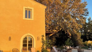 A yellow building with a large arched window and two smaller windows. There are outdoor seating areas with chairs and tables, and a large tree in the background. - Saint Rémy de Provence -mas de Charme à 3 Km du Centre (Saint-Rémy-de-Provence)
