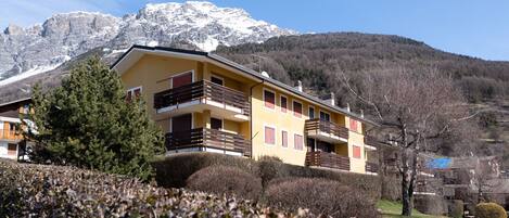 A yellow building with balconies, surrounded by trees and mountains.