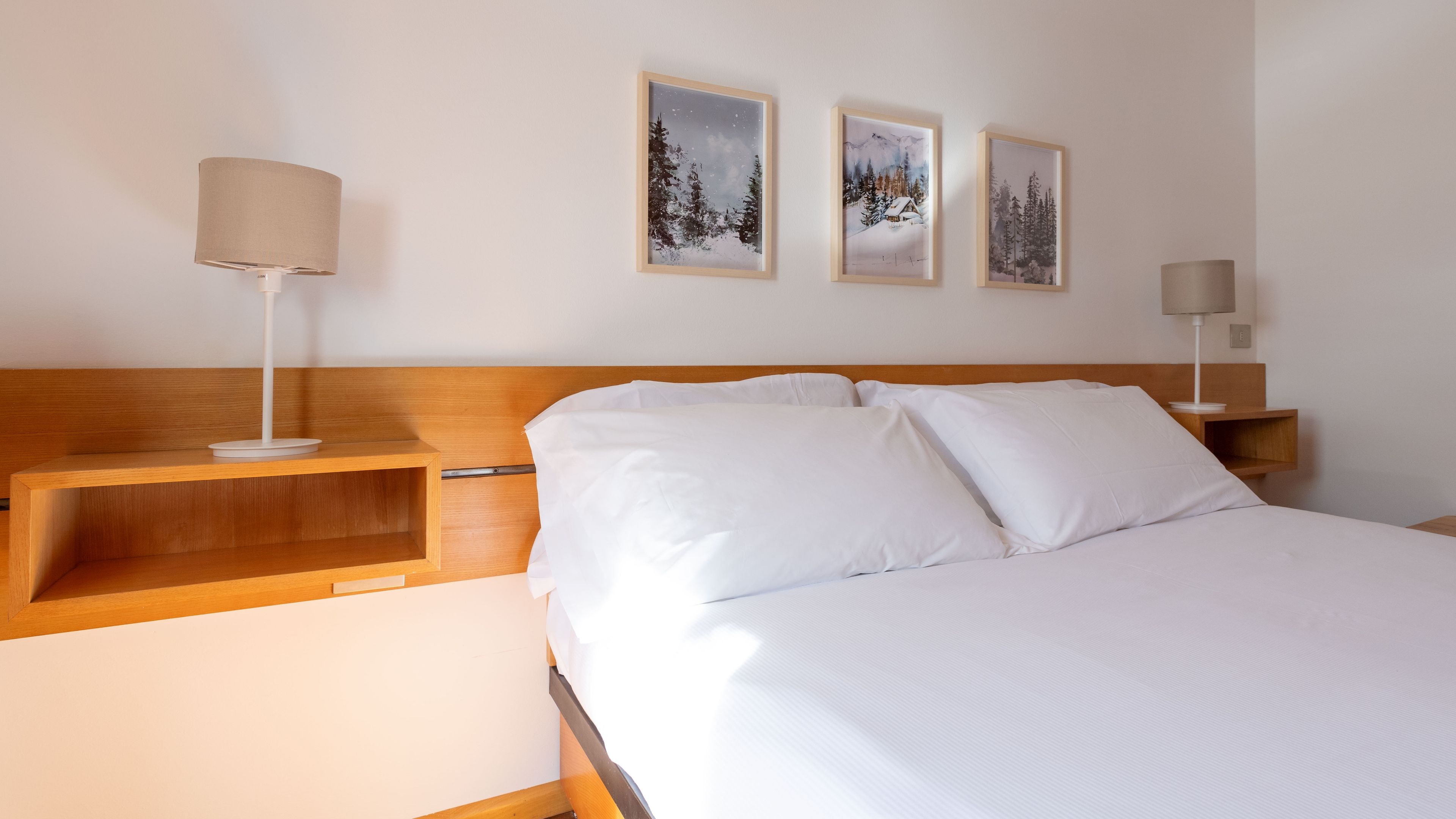 Habitación de hotel con cabecera de madera, ropa de cama blanca y dos mesitas de noche con lámparas. Hay tres cuadros en la pared.