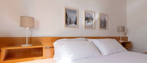 Habitación de hotel con cabecera de madera, ropa de cama blanca y dos mesitas de noche con lámparas. Hay tres cuadros en la pared.