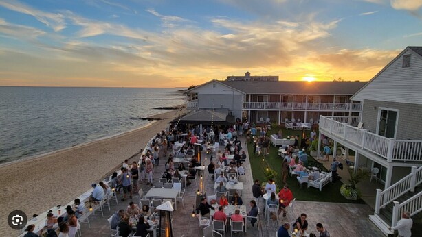Un área de comedor al aire libre frente al mar, con mesas y sillas, con vista a la playa y al océano, y un edificio al fondo.