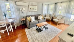 Una sala de estar con un sofá gris, una mesa de centro de madera, un tapete estampado y una zona de comedor con sillas blancas.
