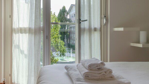 Habitación de hotel con cama, ventana con cortinas translúcidas, vista a edificios y árboles, y una pila de toallas sobre la cama.