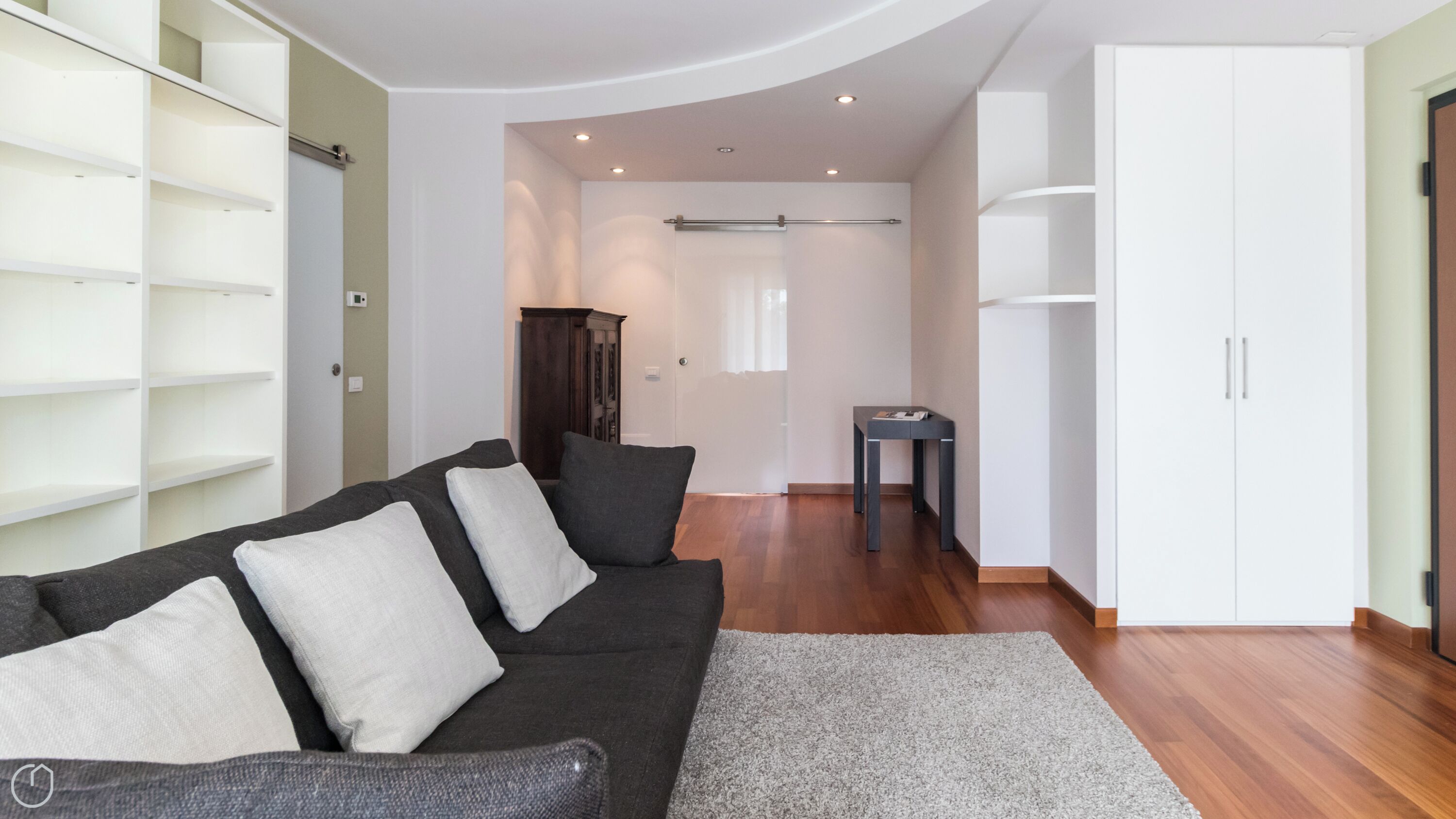 Un salon moderne avec un canapé gris foncé, des coussins blancs, un tapis gris et un parquet en bois. On y trouve une armoire blanche, une petite table et un meuble avec des étagères.