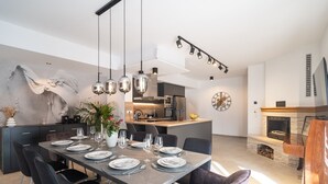 Ein moderner Essbereich mit einem langen Tisch, gedeckt für sechs Personen, umgeben von dunklen Stühlen. Im Hintergrund ist eine Küche mit Backofen und Kamin aus Stein zu sehen.