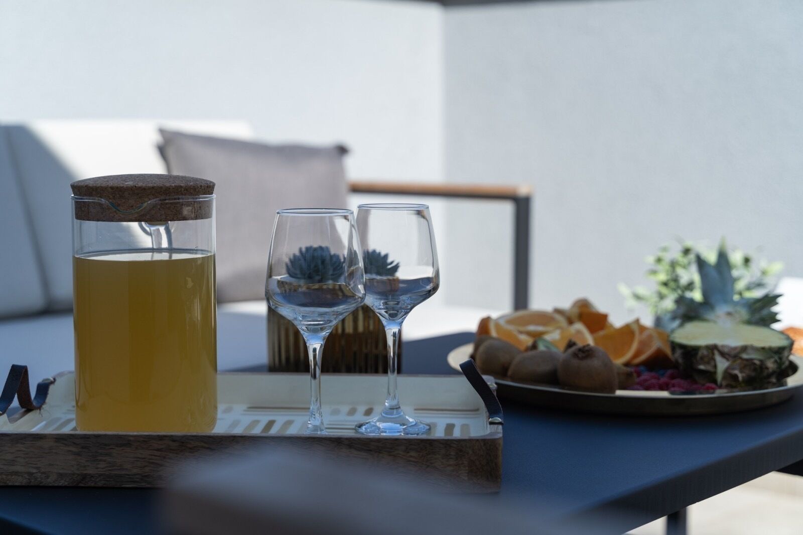 Taula amb un got de beguda groga, dos vasos de vi transparents i un plat de fruita.