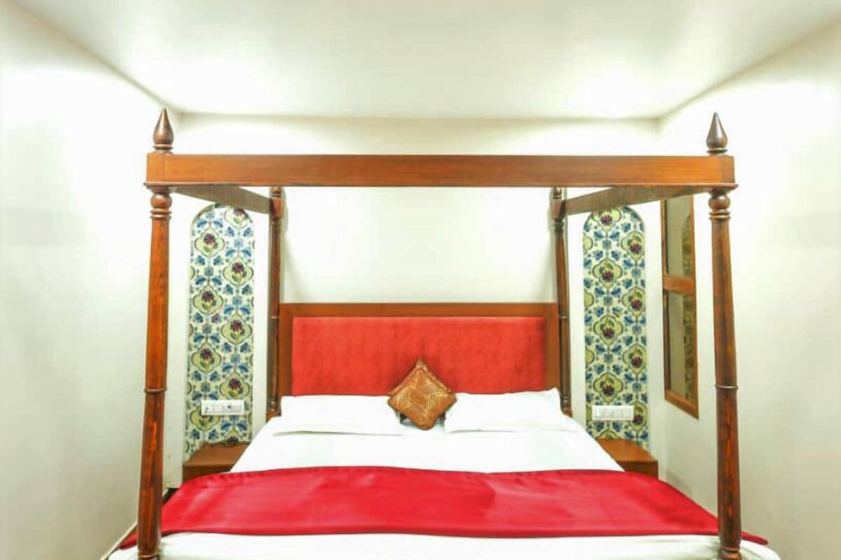 Una cama con dosel con cubrecama rojo y sábanas blancas, con papel tapiz estampado y un espejo a cada lado.