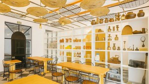 Un restaurante con mesas y sillas de madera, una pared de estanterías con diversa cerámica y utensilios de cocina, y un techo con cestas colgantes.