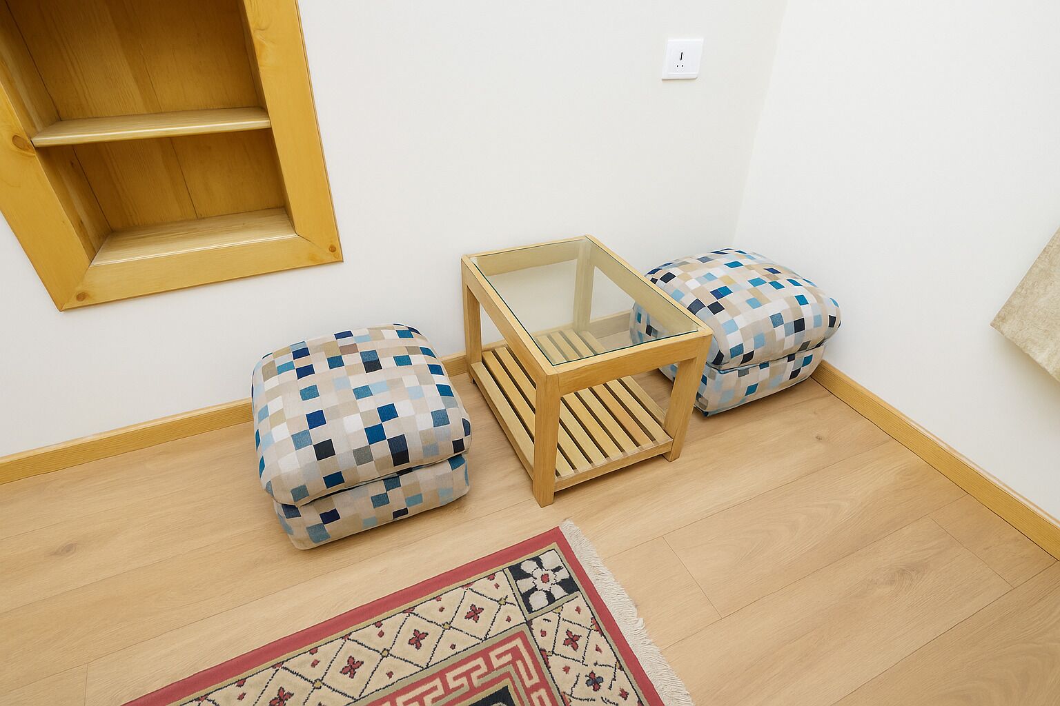 A small room with a wooden floor, a glass table, two patterned ottomans, and a patterned rug.