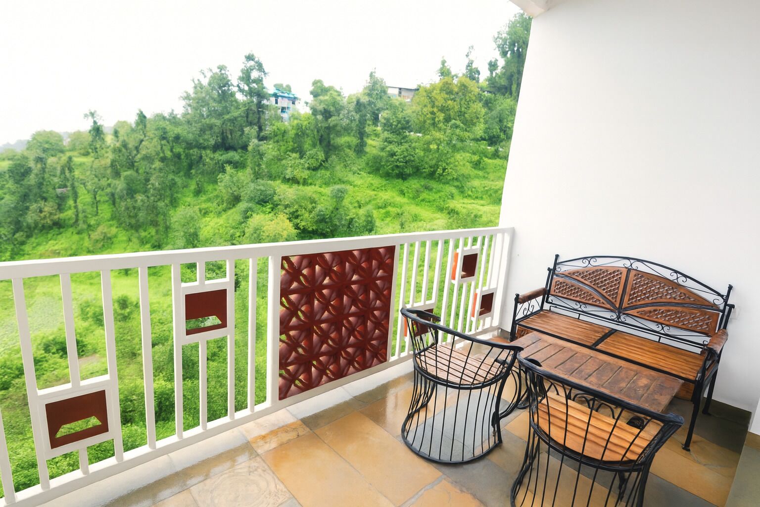 Un balcón con vista a un paisaje verde y exuberante, una mesa de madera con sillas y una baranda blanca con paneles decorativos.