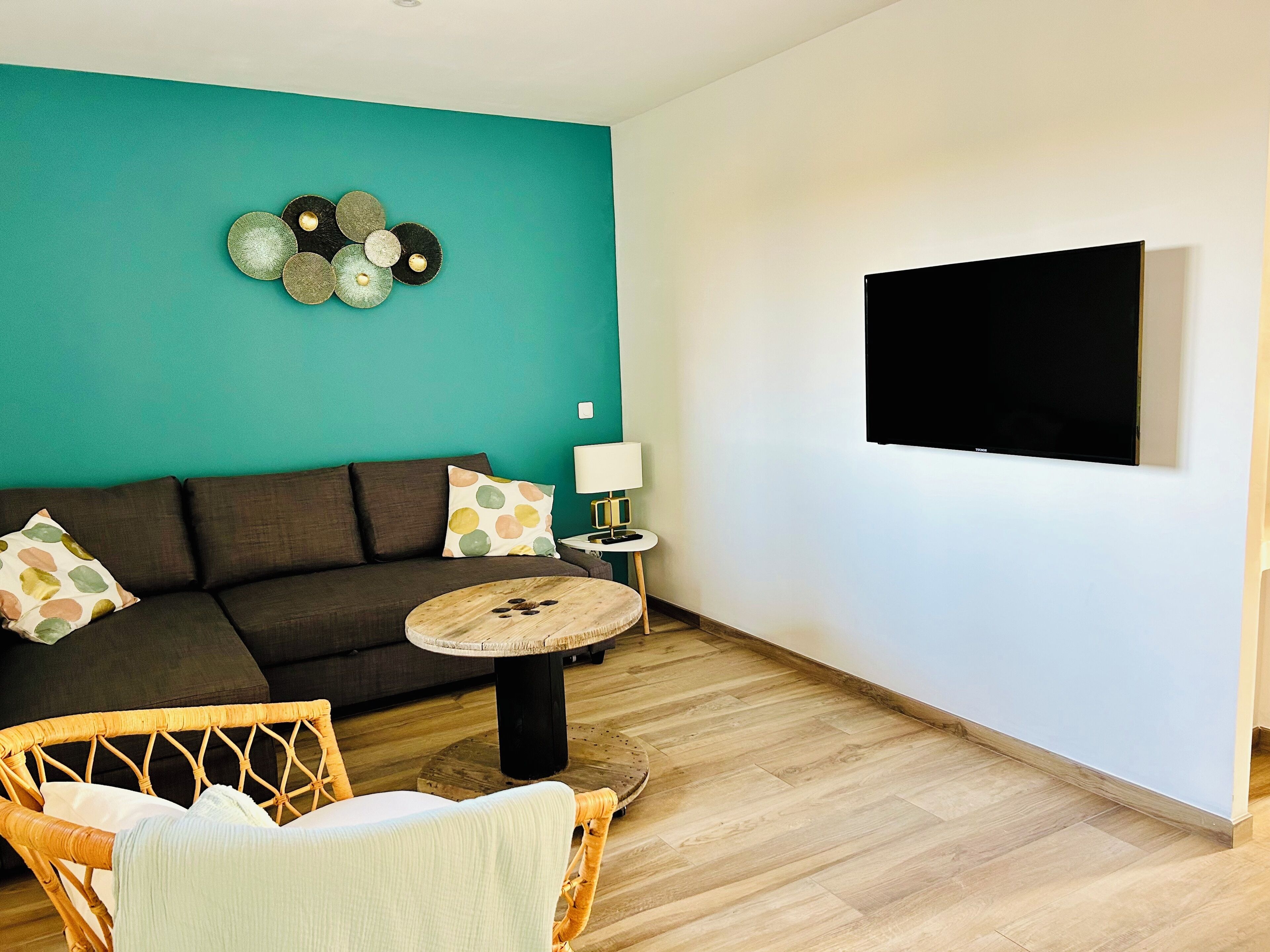 A living room with a teal wall, a dark sofa, a round coffee table, and a flat-screen TV mounted on the wall.