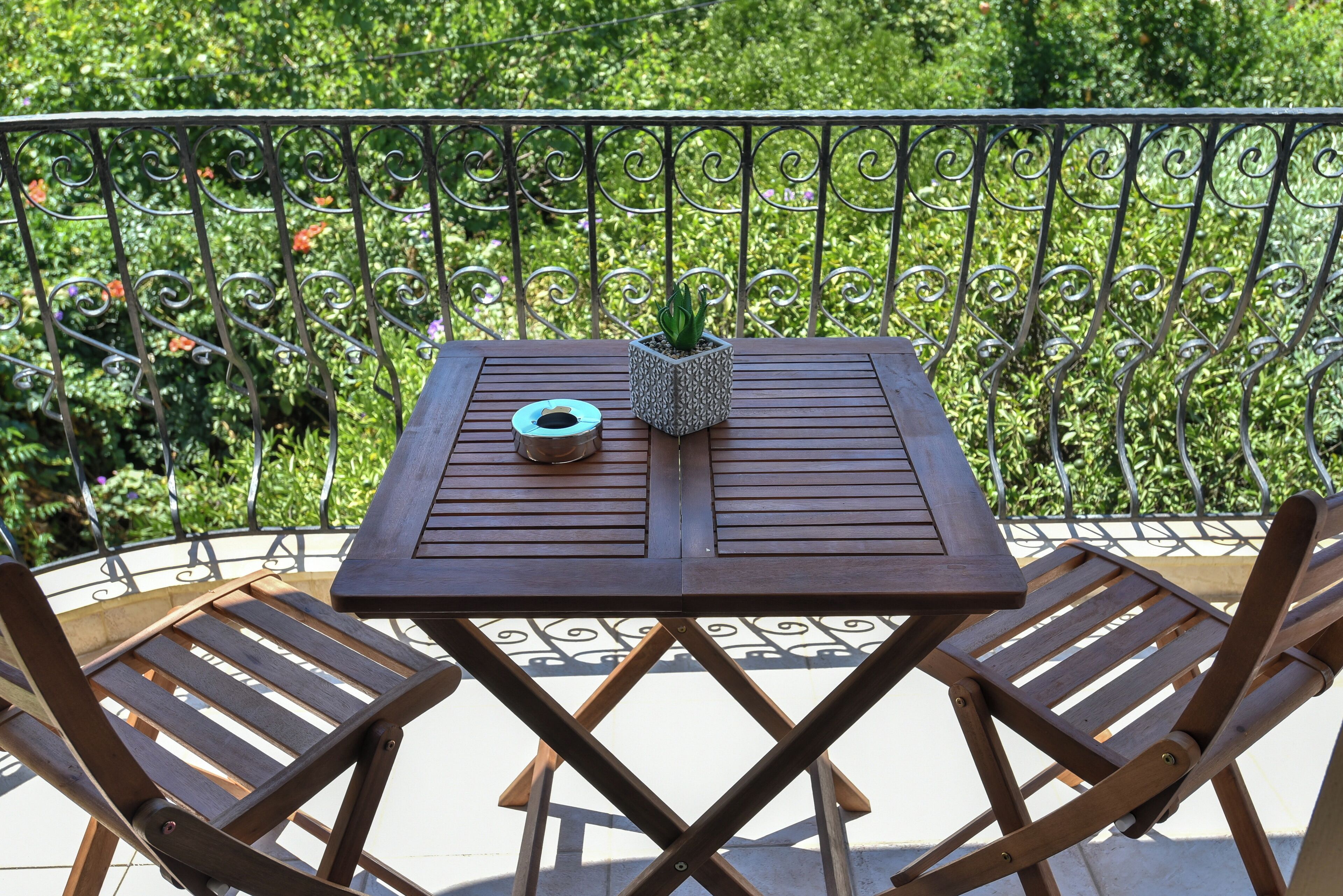 A wooden table and two chairs on an outdoor balcony with a metal railing and greenery in the background.