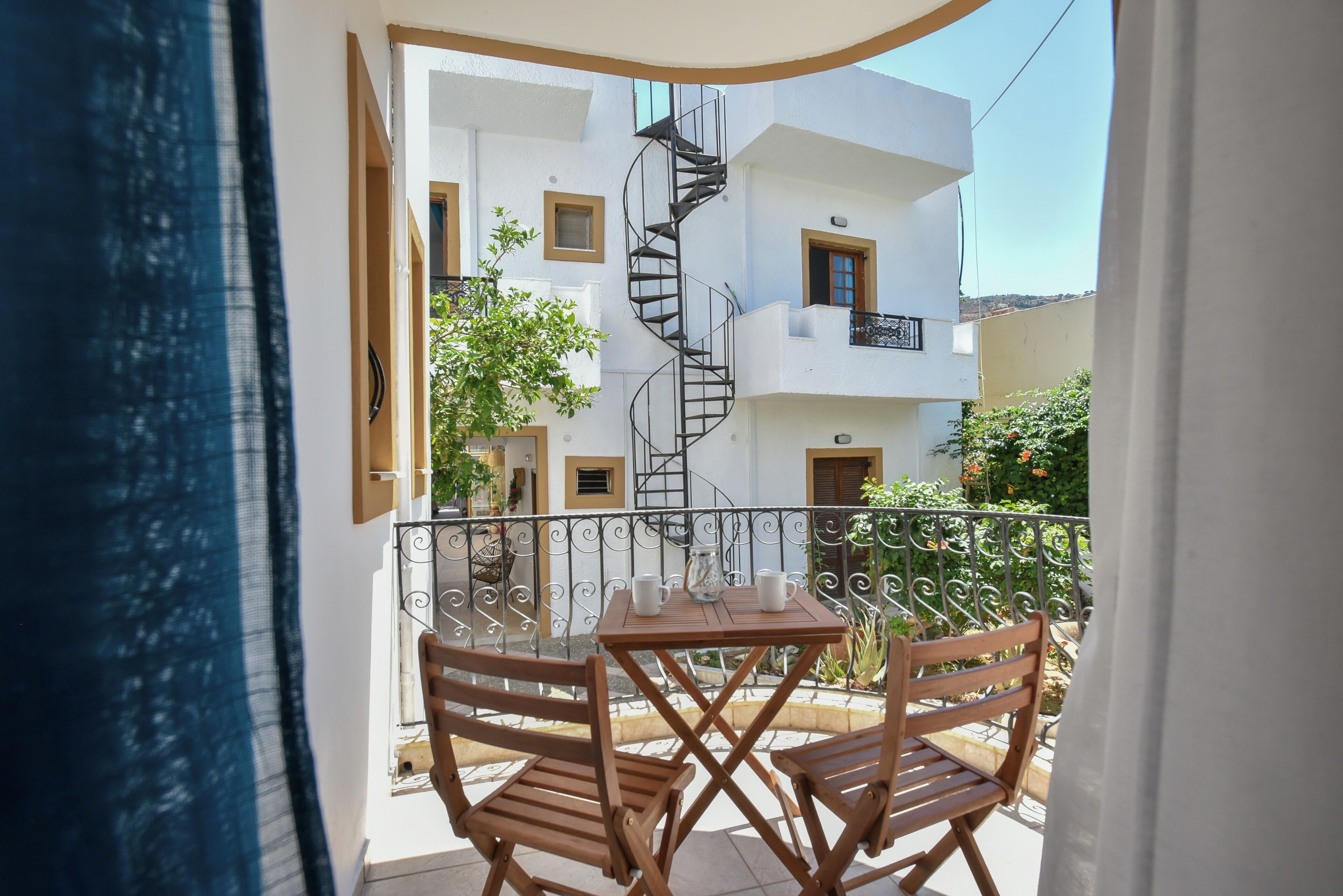 A balcony with a table and two chairs, a spiral staircase, and a white building with balconies in the background.