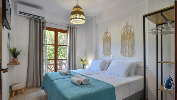 A bedroom with a bed, a hanging light fixture, a macrame wall decoration, and a window with curtains. - Eva Studios & Apartments (Malevizi)