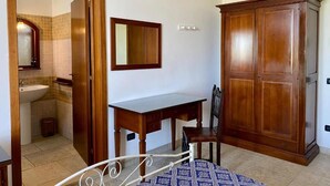 A bedroom with a bed, a wooden wardrobe, a desk, and a chair. There is a mirror on the wall and a bathroom visible through an open door.