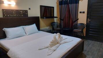 A neatly made bed with a towel arranged in a heart shape, a headboard, a wall-mounted light, a decorative panel, two chairs, a small table, and a door.