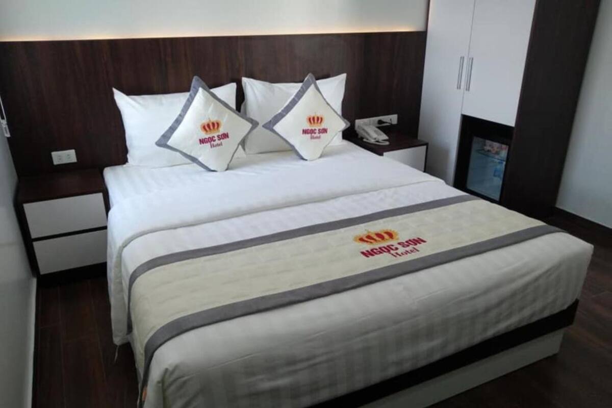 Ein Hotelzimmer mit einem großen Bett, zwei Kopfkissen mit dem Logo des Hotels, einem Nachttisch mit einem Telefon und einem Schrank.