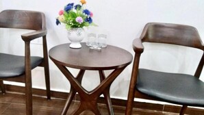 A small round table with a white vase of flowers and two glasses, flanked by two wooden chairs with grey cushions.