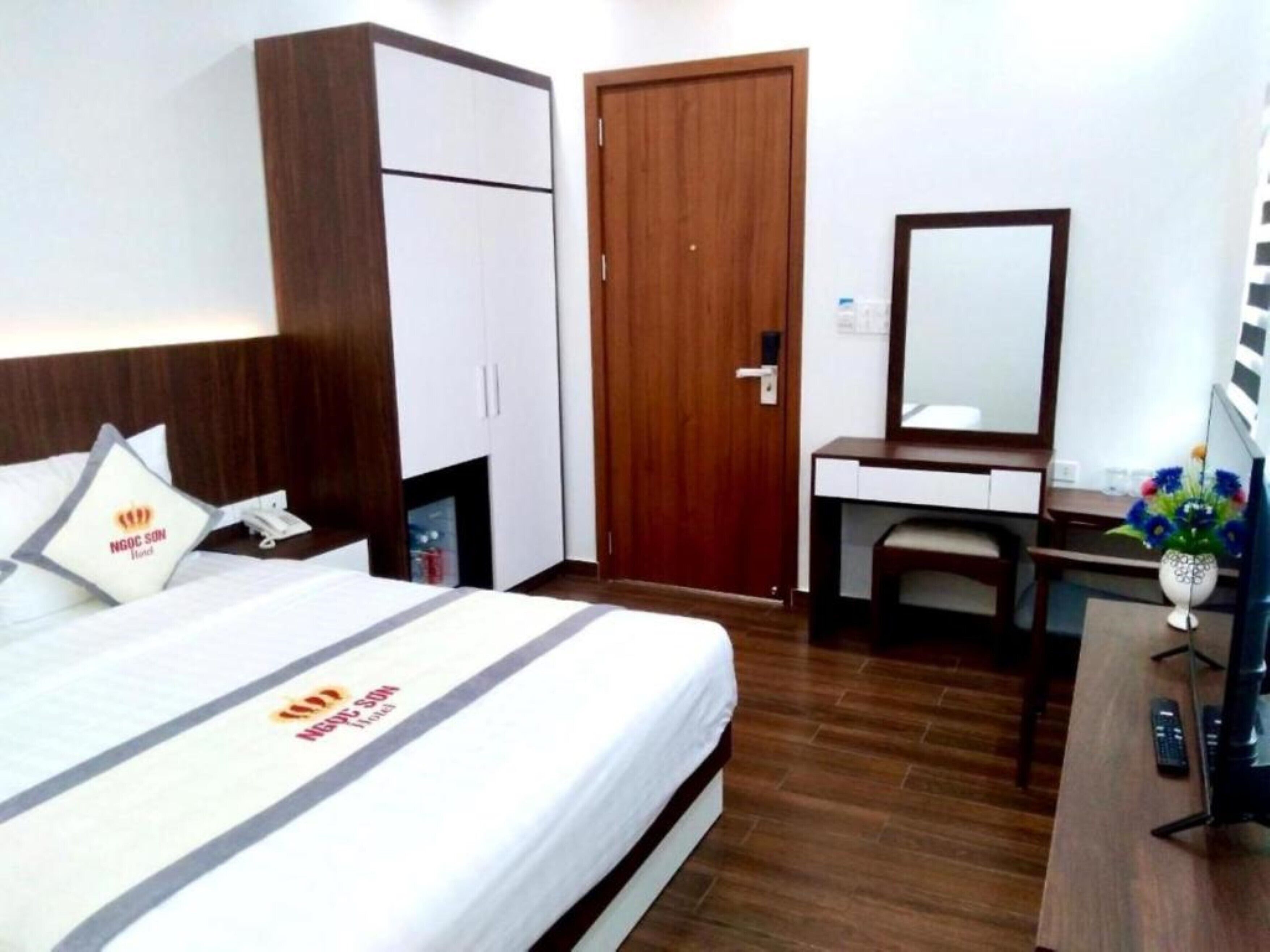 A hotel room with a bed, a wardrobe, a desk with a mirror, and a vase with flowers.