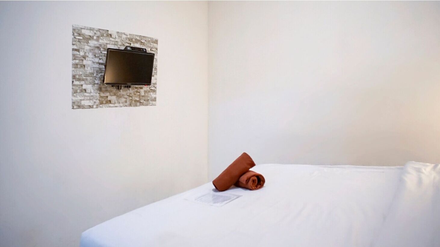 A hotel room with a flat-screen TV mounted on a textured wall, a neatly made bed with white linens, and a small folded towel on the bed.