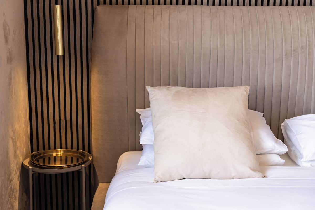 A neatly made bed with white linens and a large beige pillow, a bedside table with a metallic tray, and a wall with vertical wooden slats and a hanging light fixture.