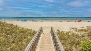 Ein hölzerner Steg führt zu einem Sandstrand mit Zelten und Menschen.